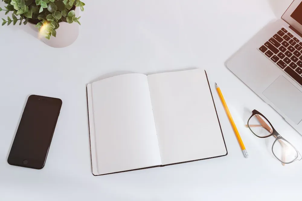 A notebook, pencil, laptop, phone and reading glasses on a table, illustrating the search for available Company Names.
